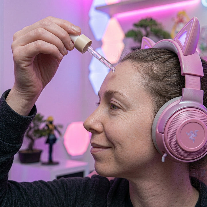 A woman wearing pink cat-ear headphones applies a clear liquid from a glass dropper to her forehead in a brightly lit room with pink ambient lighting.