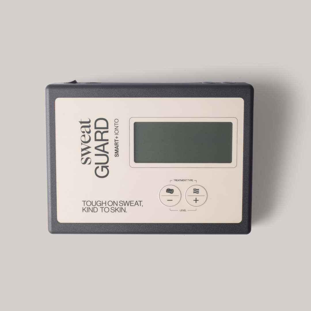 A Sweat Guard smart temperature and humidity monitor with a dark gray casing and a cream-colored front panel, featuring a digital display screen and two control buttons.