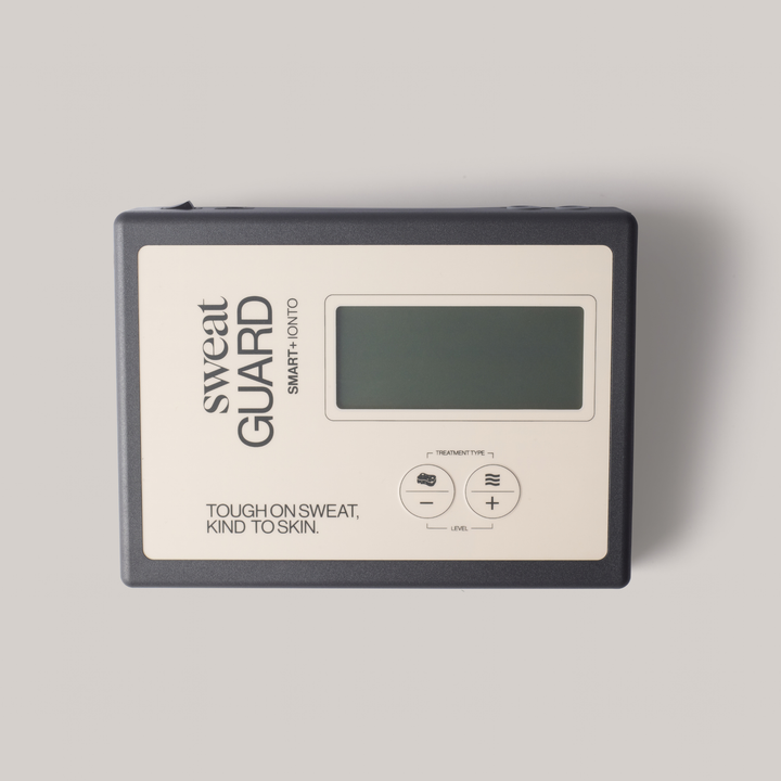 A Sweat Guard smart temperature and humidity monitor with a dark gray casing and a cream-colored front panel, featuring a digital display screen and two control buttons.