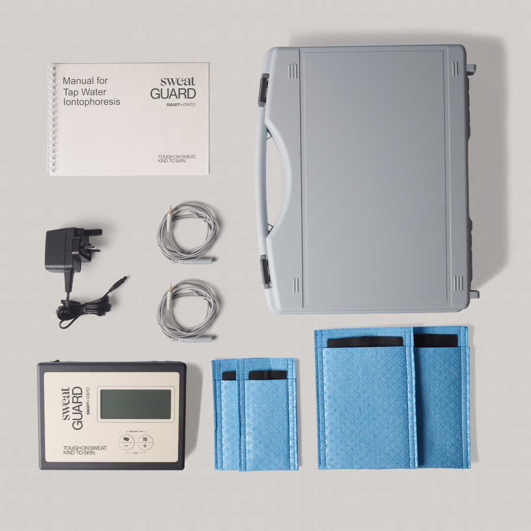 Sweat Guard iontophoresis kit laid out on a light gray background, including a gray carrying case, control unit, power adapter, cables, blue pouches, and a printed user manual.